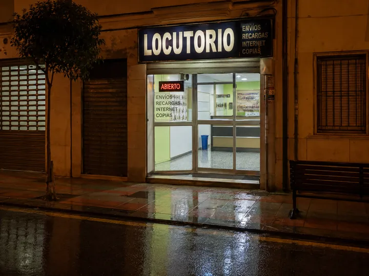 Fachada de locutorio de noche con lluvia, reflejos en el asfalto